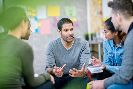 A diverse group of young professionals sits in a circle, engaged in a collaborative discussion in a modern office setting.