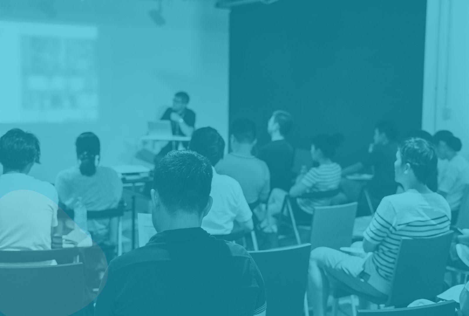 A group of students sits in a classroom listening to a teacher who is presenting at the front of the room. The entire image is overlaid with a blue tint.